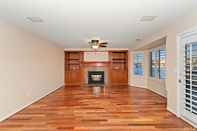 unfurnished living room featuring a tiled fireplace, ceiling fan, light wood-type flooring, and built in shelves