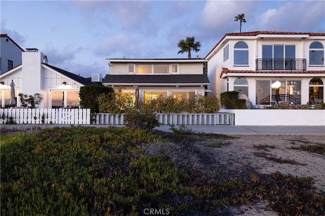 back of property at dusk featuring a balcony, a fenced front yard, a tile roof, and stucco siding