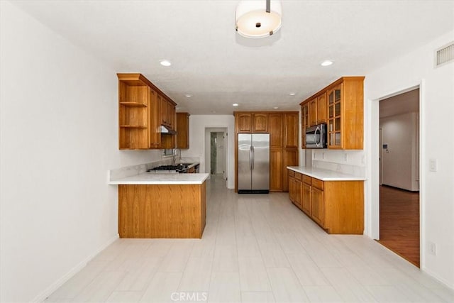 kitchen with stainless steel appliances, light countertops, open shelves, brown cabinetry, and glass insert cabinets