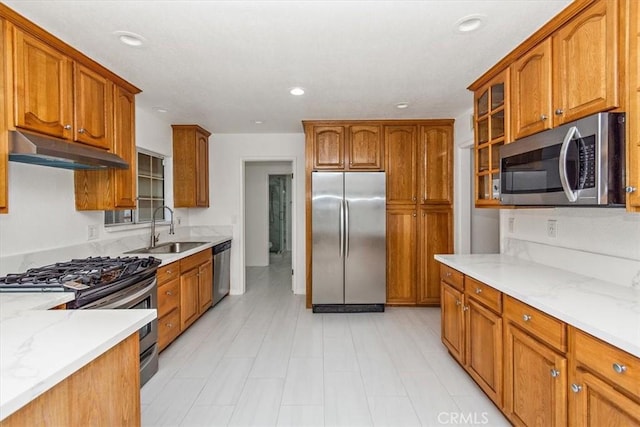 kitchen featuring under cabinet range hood, a sink, appliances with stainless steel finishes, brown cabinetry, and glass insert cabinets