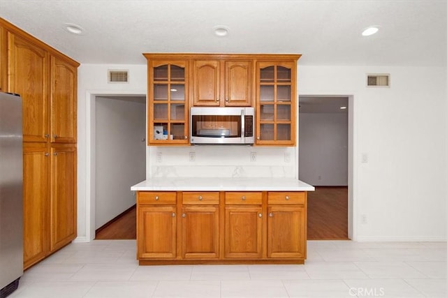 kitchen featuring visible vents, light countertops, appliances with stainless steel finishes, brown cabinetry, and glass insert cabinets