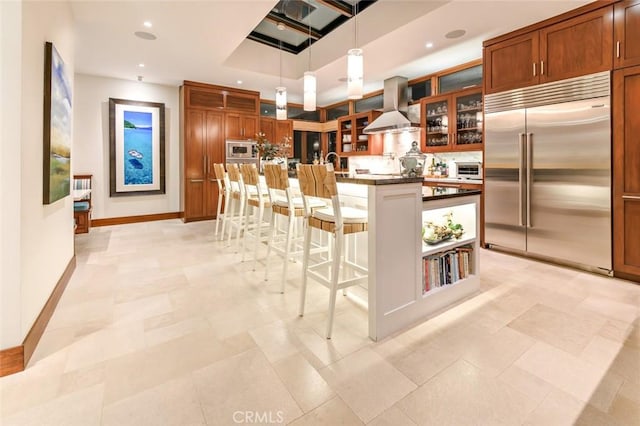 kitchen featuring a center island with sink, built in appliances, a kitchen breakfast bar, decorative light fixtures, and wall chimney range hood