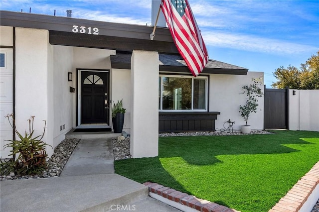 view of exterior entry featuring stucco siding, roof with shingles, fence, and a yard