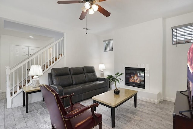 living area featuring a ceiling fan, baseboards, stairway, light wood finished floors, and a glass covered fireplace