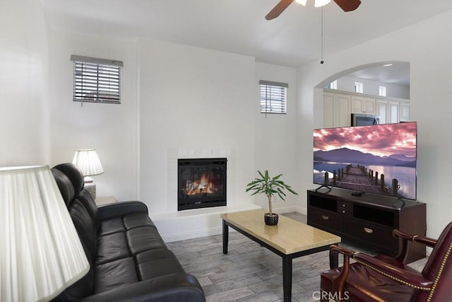 living room with arched walkways, light wood-style flooring, a glass covered fireplace, ceiling fan, and baseboards