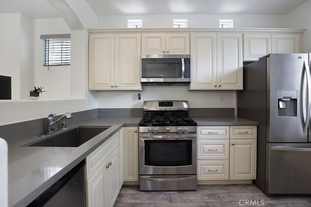 kitchen with stainless steel appliances, dark countertops, cream cabinetry, and a sink