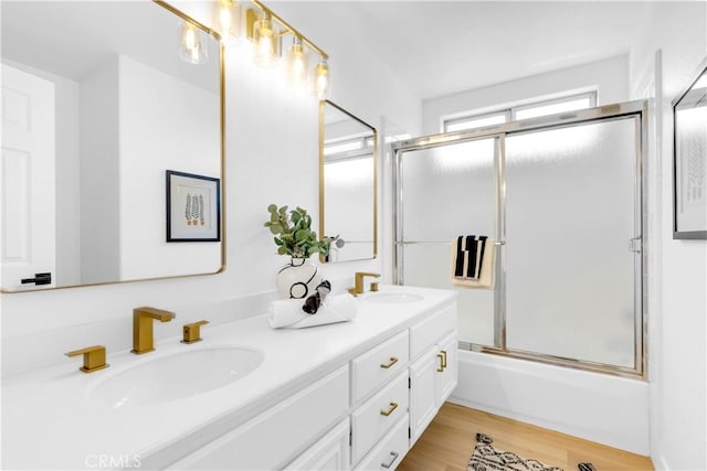 full bath with double vanity, enclosed tub / shower combo, a sink, and wood finished floors