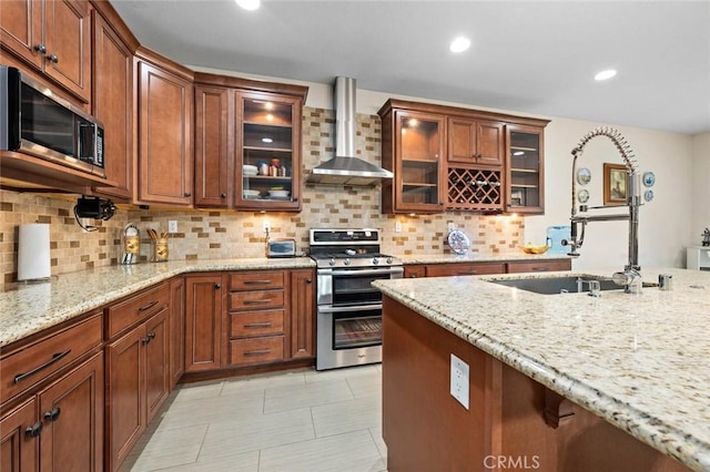 kitchen featuring light stone counters, stainless steel appliances, wall chimney range hood, tasteful backsplash, and glass insert cabinets