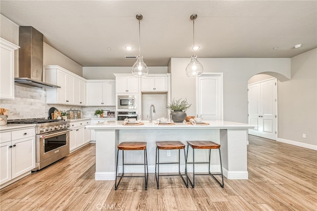 kitchen with appliances with stainless steel finishes, pendant lighting, white cabinets, a center island with sink, and wall chimney exhaust hood