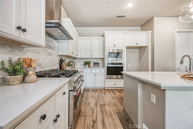 kitchen with wall chimney range hood, light hardwood / wood-style flooring, appliances with stainless steel finishes, white cabinetry, and decorative light fixtures