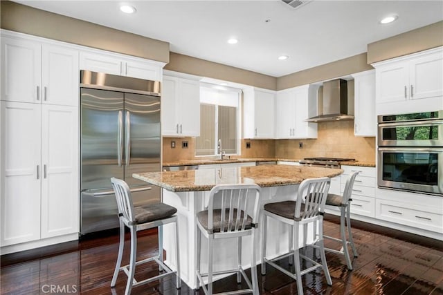 kitchen featuring light stone counters, a center island, stainless steel appliances, white cabinetry, and wall chimney exhaust hood