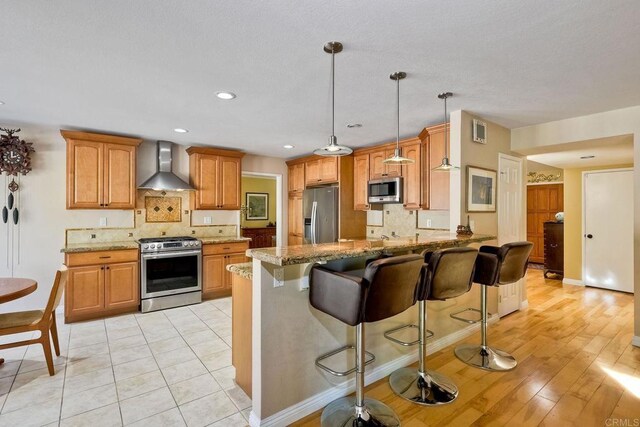 kitchen featuring light stone counters, a peninsula, stainless steel appliances, and wall chimney range hood