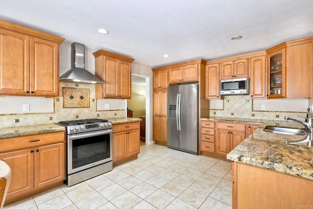 kitchen with light stone counters, appliances with stainless steel finishes, wall chimney exhaust hood, and a sink