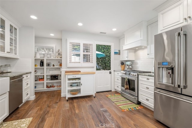 kitchen with glass insert cabinets, light stone counters, custom exhaust hood, stainless steel appliances, and white cabinetry