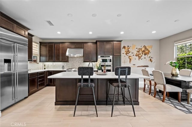 kitchen featuring stainless steel appliances, light countertops, a kitchen island with sink, dark brown cabinetry, and under cabinet range hood