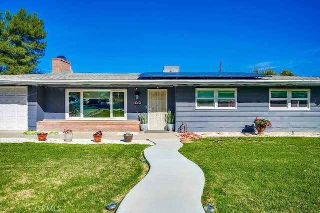 ranch-style home with a front yard, a chimney, an attached garage, roof mounted solar panels, and brick siding