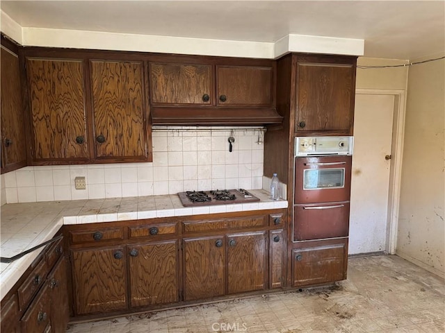 kitchen with tile countertops, oven, under cabinet range hood, a warming drawer, and stainless steel gas stovetop