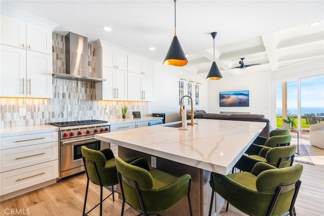 kitchen with high end stove, a sink, white cabinets, wall chimney exhaust hood, and an island with sink