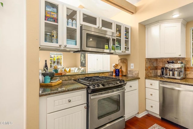 kitchen with tasteful backsplash, dark stone counters, glass insert cabinets, stainless steel appliances, and white cabinetry