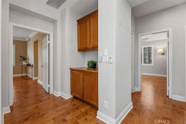 hall with light wood-style flooring, baseboards, visible vents, and ornamental molding