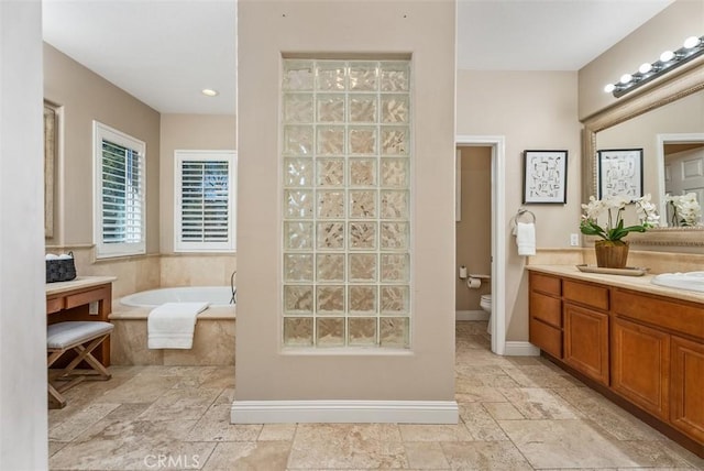 bathroom with toilet, stone tile floors, double vanity, baseboards, and a bath