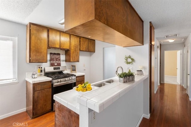 kitchen with sink, tile counters, kitchen peninsula, stainless steel gas range oven, and light hardwood / wood-style flooring