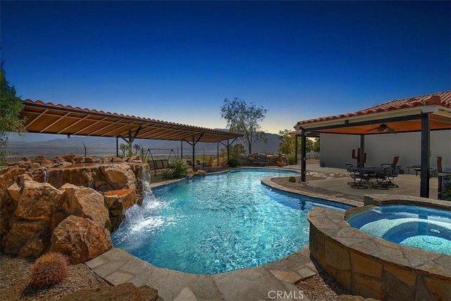 pool at dusk featuring a patio area, pool water feature, and an in ground hot tub