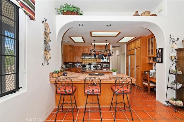 kitchen with light tile patterned flooring, a breakfast bar area, tile counters, kitchen peninsula, and exhaust hood