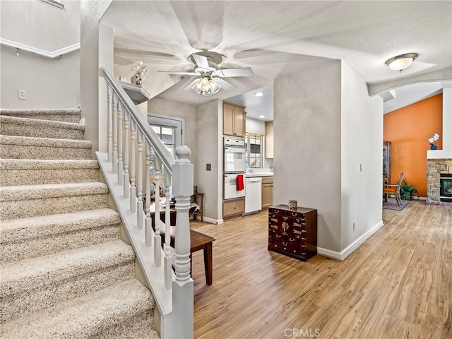 stairs featuring wood finished floors, baseboards, a ceiling fan, arched walkways, and a stone fireplace