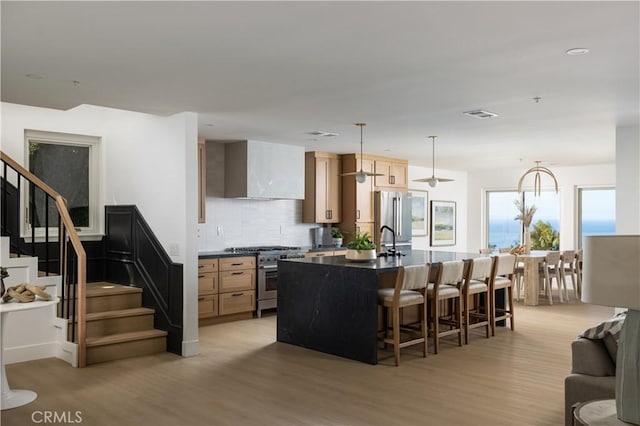 kitchen featuring high quality appliances, a center island with sink, a kitchen breakfast bar, dark countertops, and wall chimney range hood