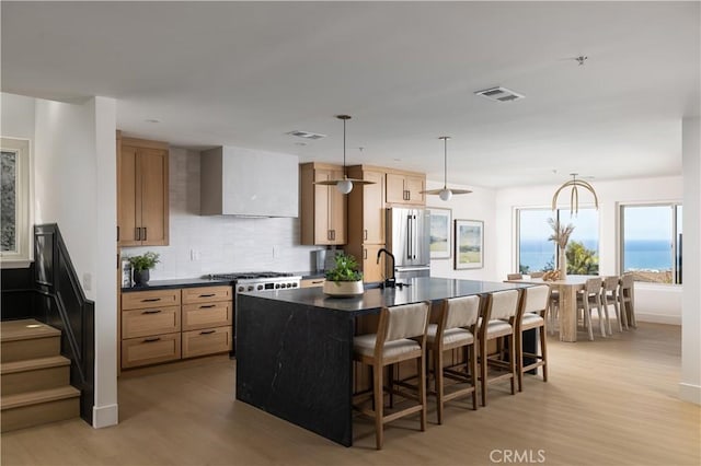 kitchen featuring dark countertops, a breakfast bar area, an island with sink, appliances with stainless steel finishes, and wall chimney exhaust hood