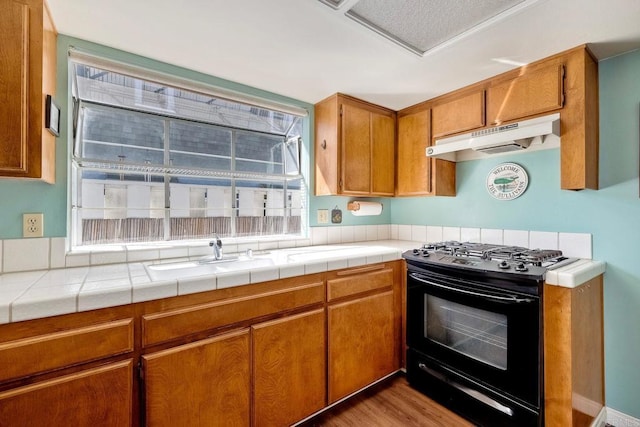 kitchen featuring tile counters, brown cabinets, under cabinet range hood, a sink, and gas stove