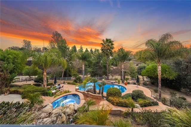 pool at dusk with fence, a hot tub, a community pool, and a patio