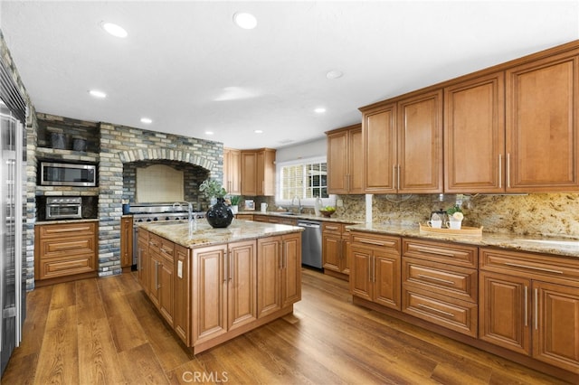 kitchen featuring wood finished floors, appliances with stainless steel finishes, brown cabinets, light stone countertops, and tasteful backsplash