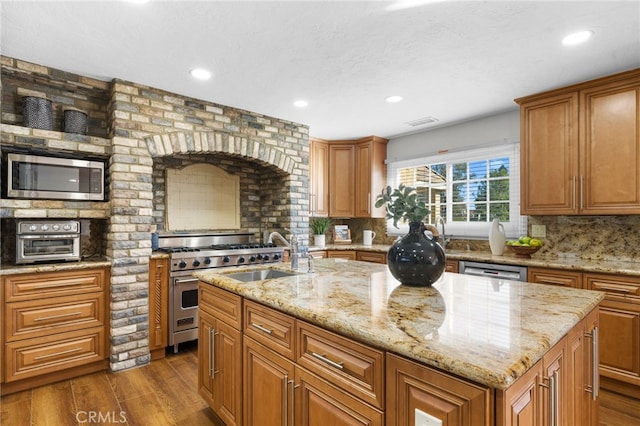 kitchen featuring a sink, appliances with stainless steel finishes, backsplash, dark wood finished floors, and a center island with sink