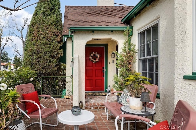 doorway to property featuring a patio, a shingled roof, a chimney, and stucco siding