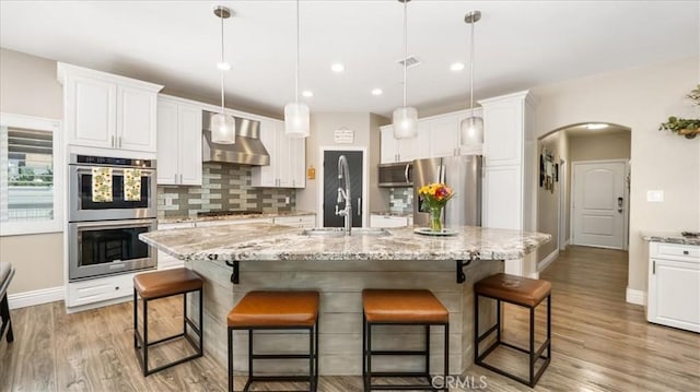 kitchen featuring arched walkways, stainless steel appliances, white cabinets, a sink, and wall chimney range hood