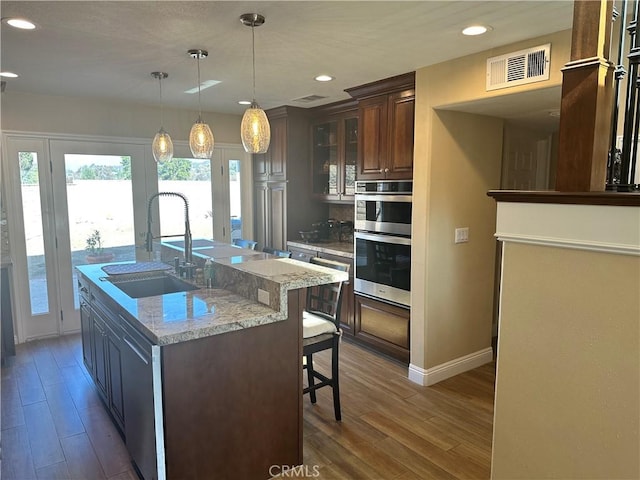 kitchen with an island with sink, hanging light fixtures, stainless steel double oven, light stone countertops, and sink