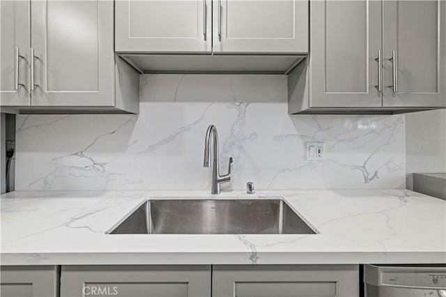 kitchen featuring gray cabinets, a sink, light stone counters, backsplash, and stainless steel dishwasher