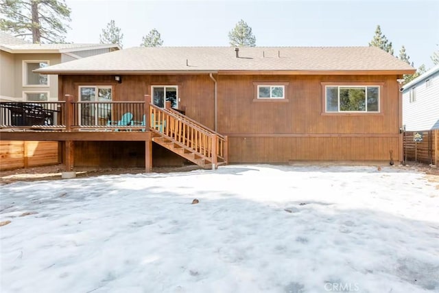 snow covered rear of property with a wooden deck, fence, and roof with shingles