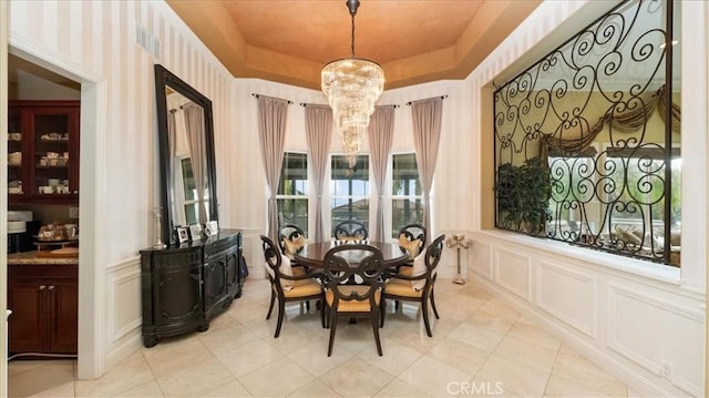 dining area with a tray ceiling, a decorative wall, wainscoting, light tile patterned flooring, and a chandelier