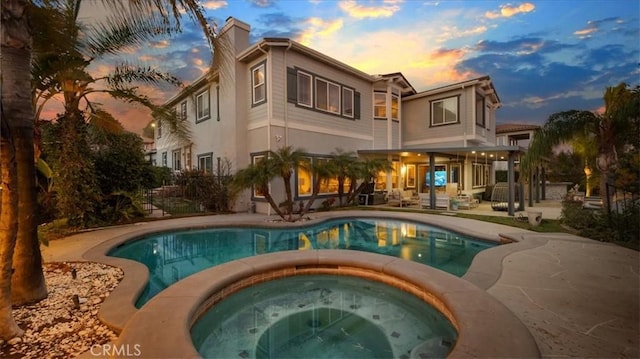 back of house at dusk featuring an in ground hot tub, a chimney, a patio area, and an outdoor pool