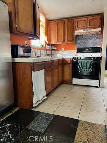 kitchen featuring under cabinet range hood, range with gas stovetop, freestanding refrigerator, brown cabinetry, and black microwave