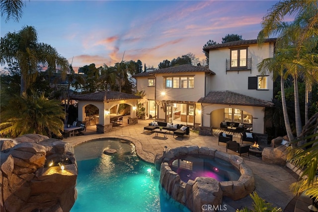 back house at dusk with a swimming pool with hot tub, a balcony, a patio area, exterior kitchen, and an outdoor living space with a fire pit