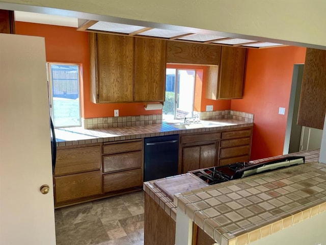 kitchen with brown cabinetry, dishwasher, tile countertops, freestanding refrigerator, and a sink