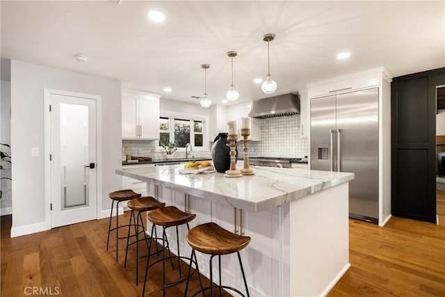 kitchen with built in fridge, a center island, white cabinets, wood finished floors, and wall chimney exhaust hood