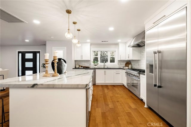 kitchen featuring high end appliances, a sink, wall chimney range hood, a kitchen island, and light wood-type flooring