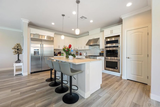 kitchen featuring built in appliances, an island with sink, pendant lighting, sink, and white cabinetry