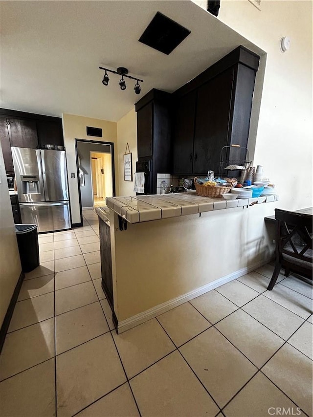 kitchen with stainless steel fridge with ice dispenser, tile countertops, light tile patterned floors, a peninsula, and dark cabinetry