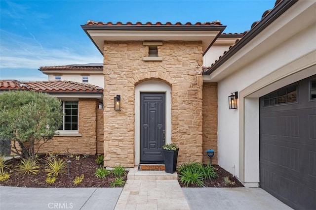 doorway to property with stone siding, a tile roof, an attached garage, roof mounted solar panels, and stucco siding